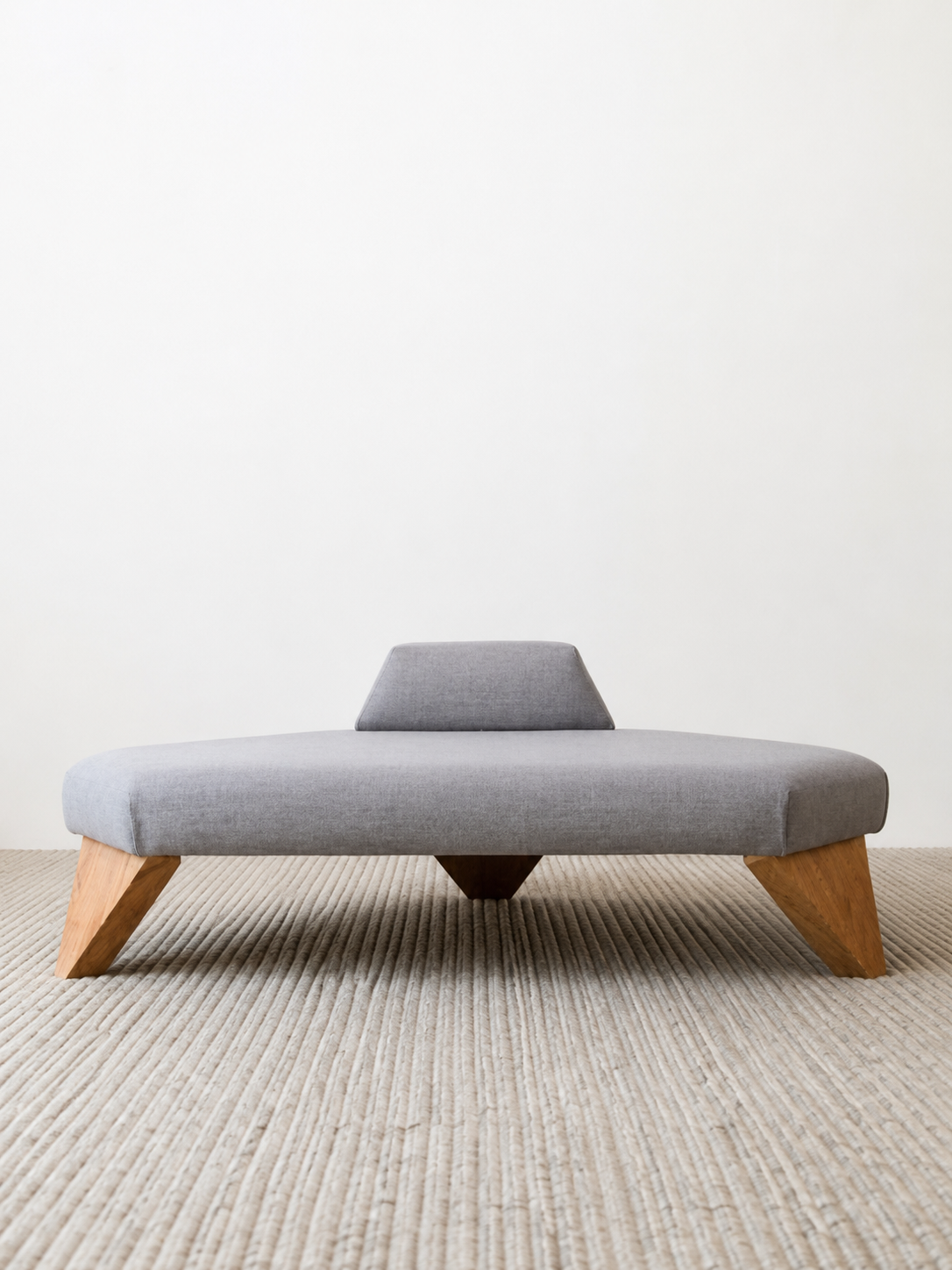 Segunda versión del banco de meditación: pieza baja tapizada en gris con respaldo en cuña y patas de madera, vista frontal