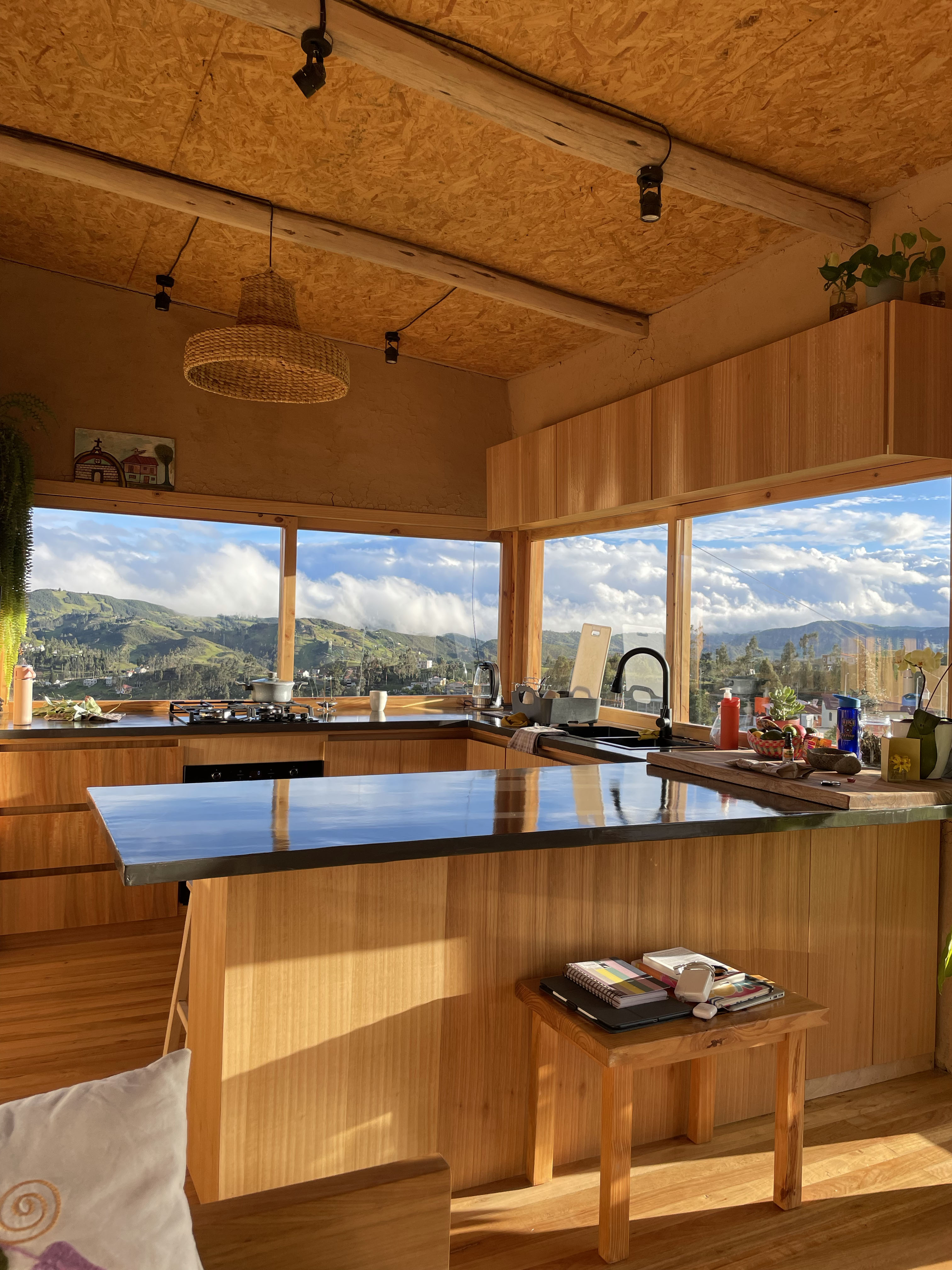 Casa Bhumi — cocina en U con mesones de concreto negro y muebles de madera, ventanas en esquina hacia el valle, cielo raso de tablero OSB