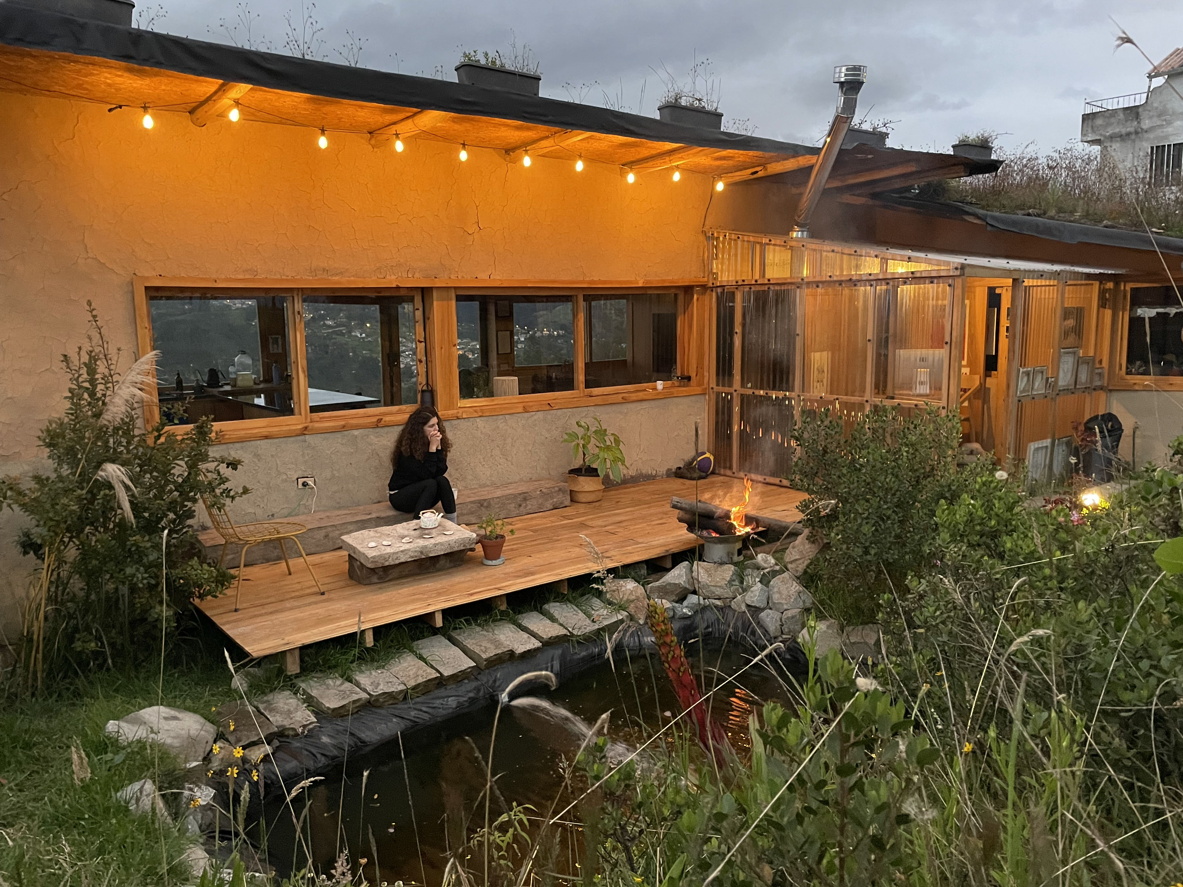 Casa Bhumi — deck de madera junto a un estanque, mesa de té de piedra y fogón al anochecer, muros de tierra iluminados por guirnaldas