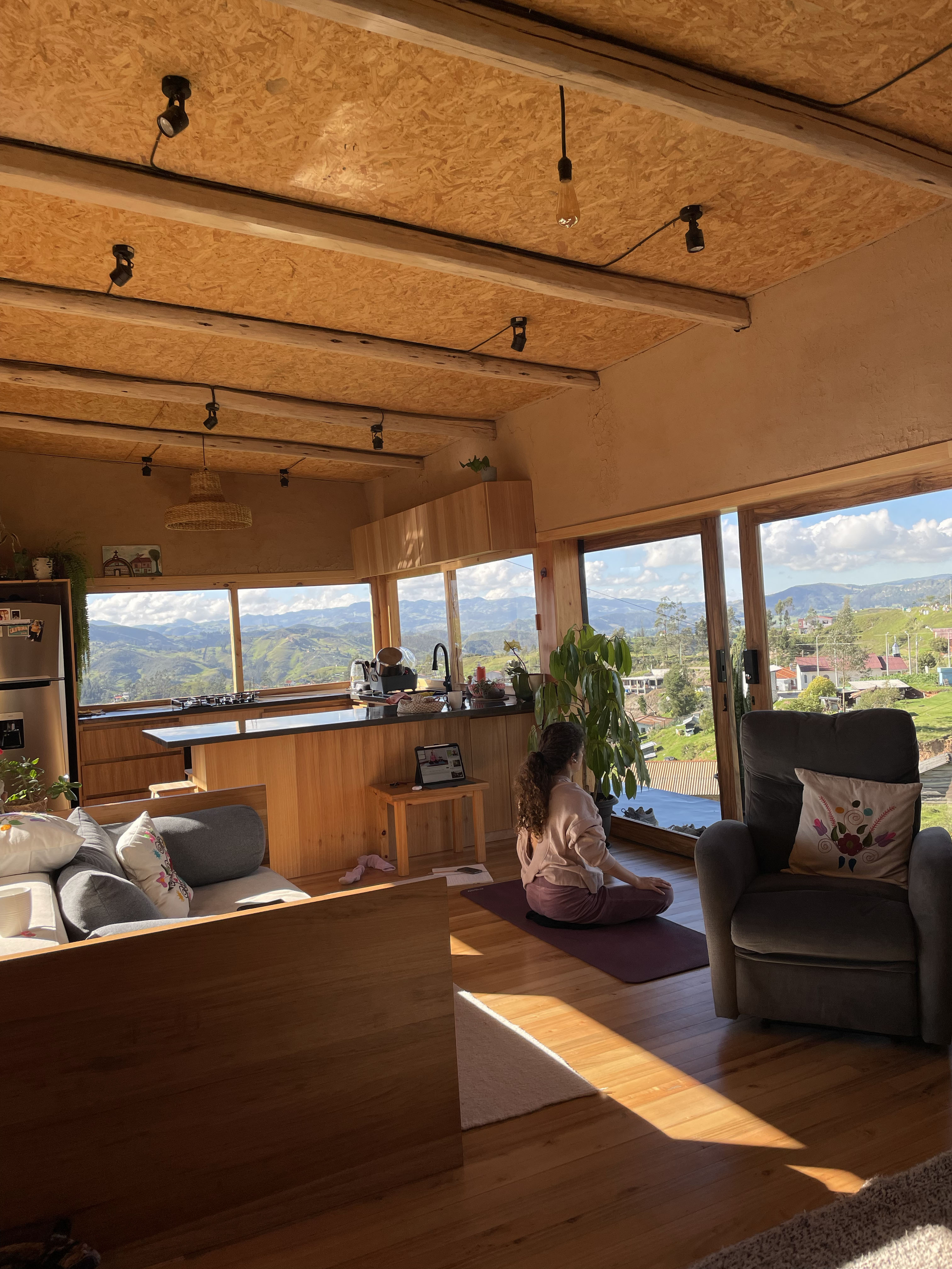 Casa Bhumi — estar con piso de madera y gran ventanal corredizo al valle, luz de la tarde entrando