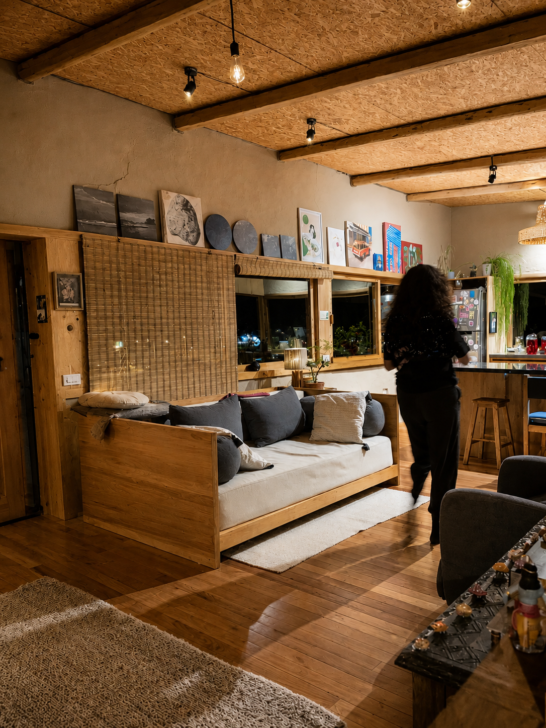 Casa Bhumi — estar de noche con banca-cama de madera, celosía de caña y repisa de arte sobre los muros de tierra