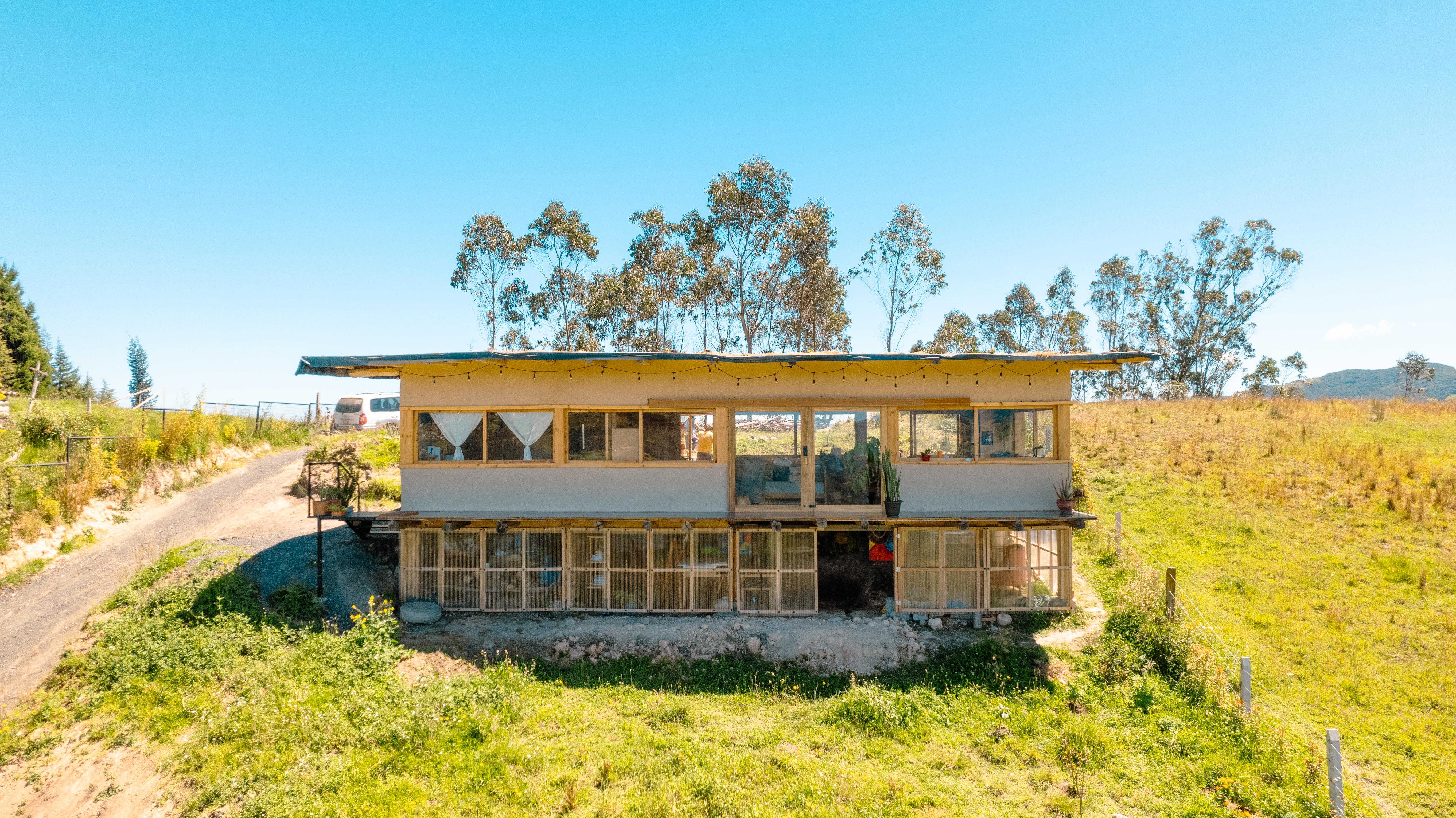 Casa Bhumi — fachada frontal de dos niveles: la planta de vivienda sobre un zócalo de invernadero cerrado con caña y policarbonato