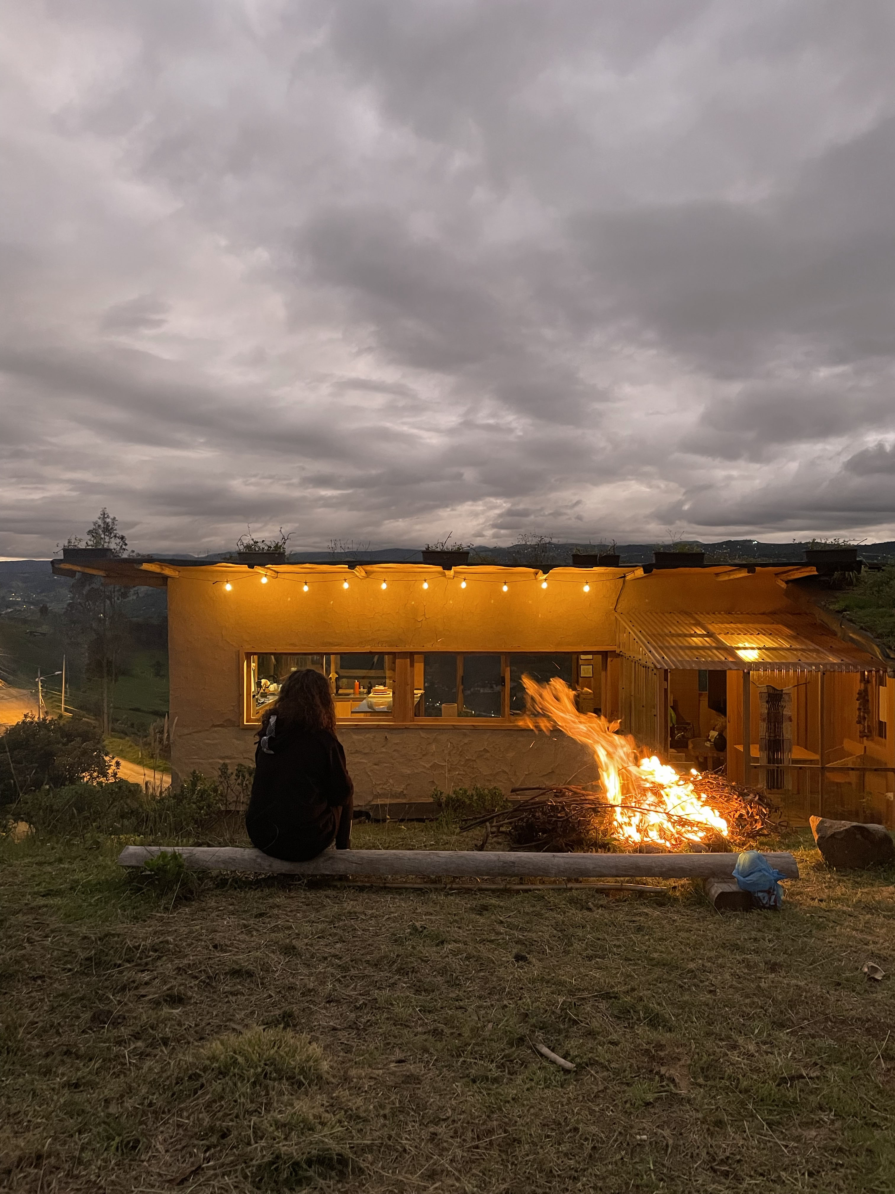 Casa Bhumi — fogata frente a la casa al anochecer, muros de tierra encendidos y cubierta verde con maceteros bajo un cielo nublado
