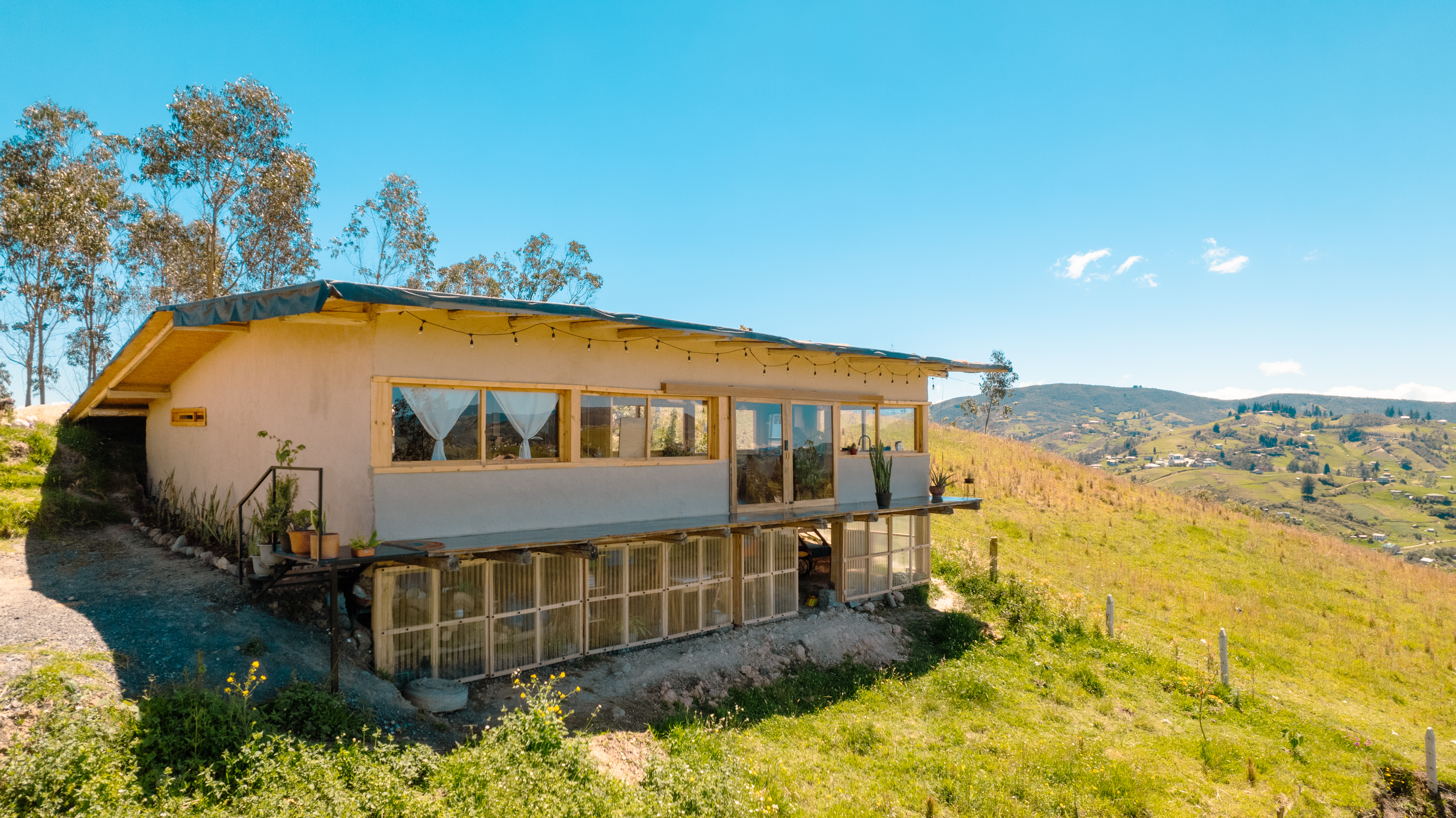 Casa Bhumi — vivienda de tierra posada sobre la ladera, abierta al valle, con cubierta verde y zócalo de invernadero