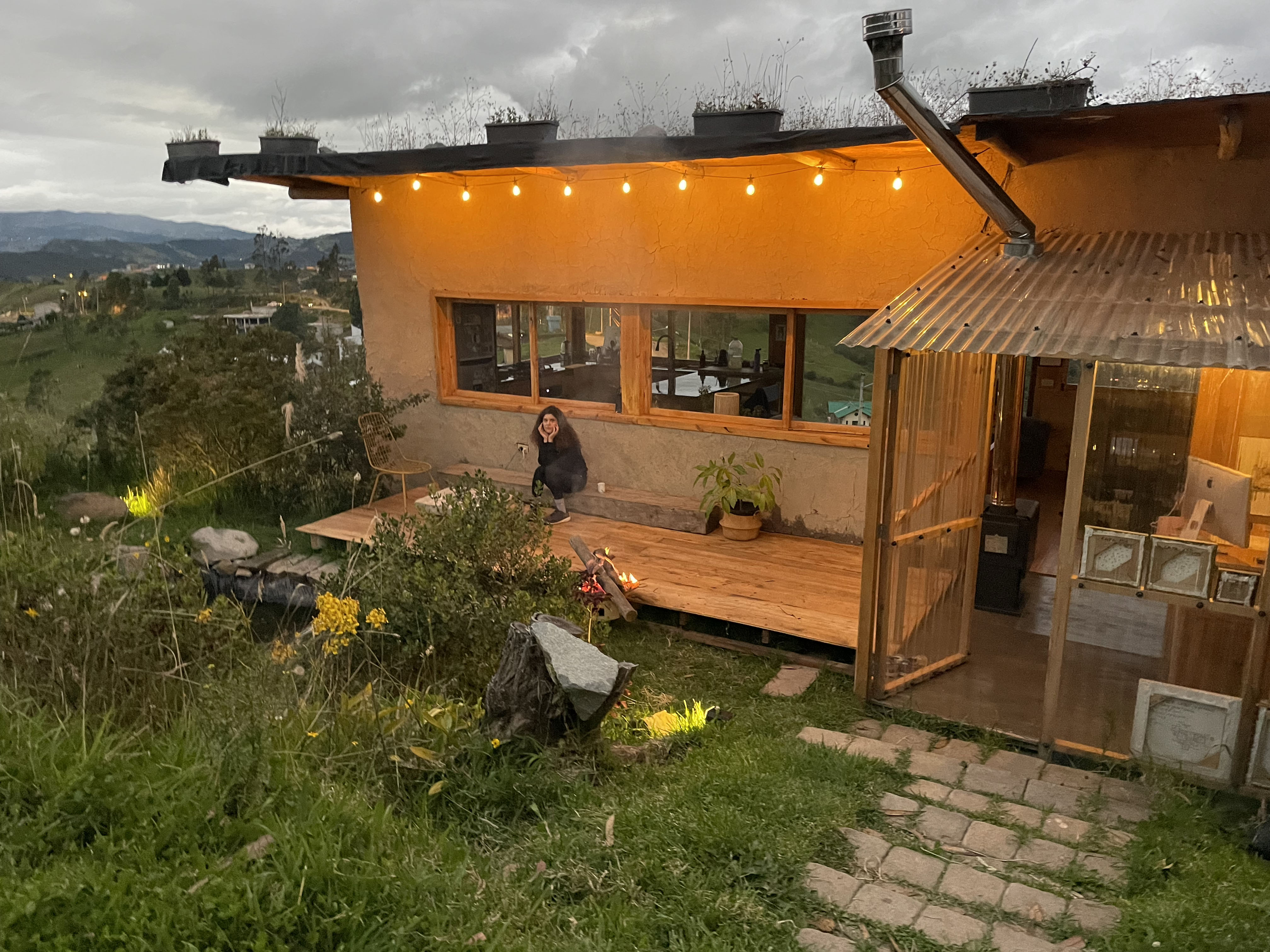 Casa Bhumi — patio al anochecer con deck de madera, maceteros sobre la cubierta verde y la galería de policarbonato iluminada por dentro