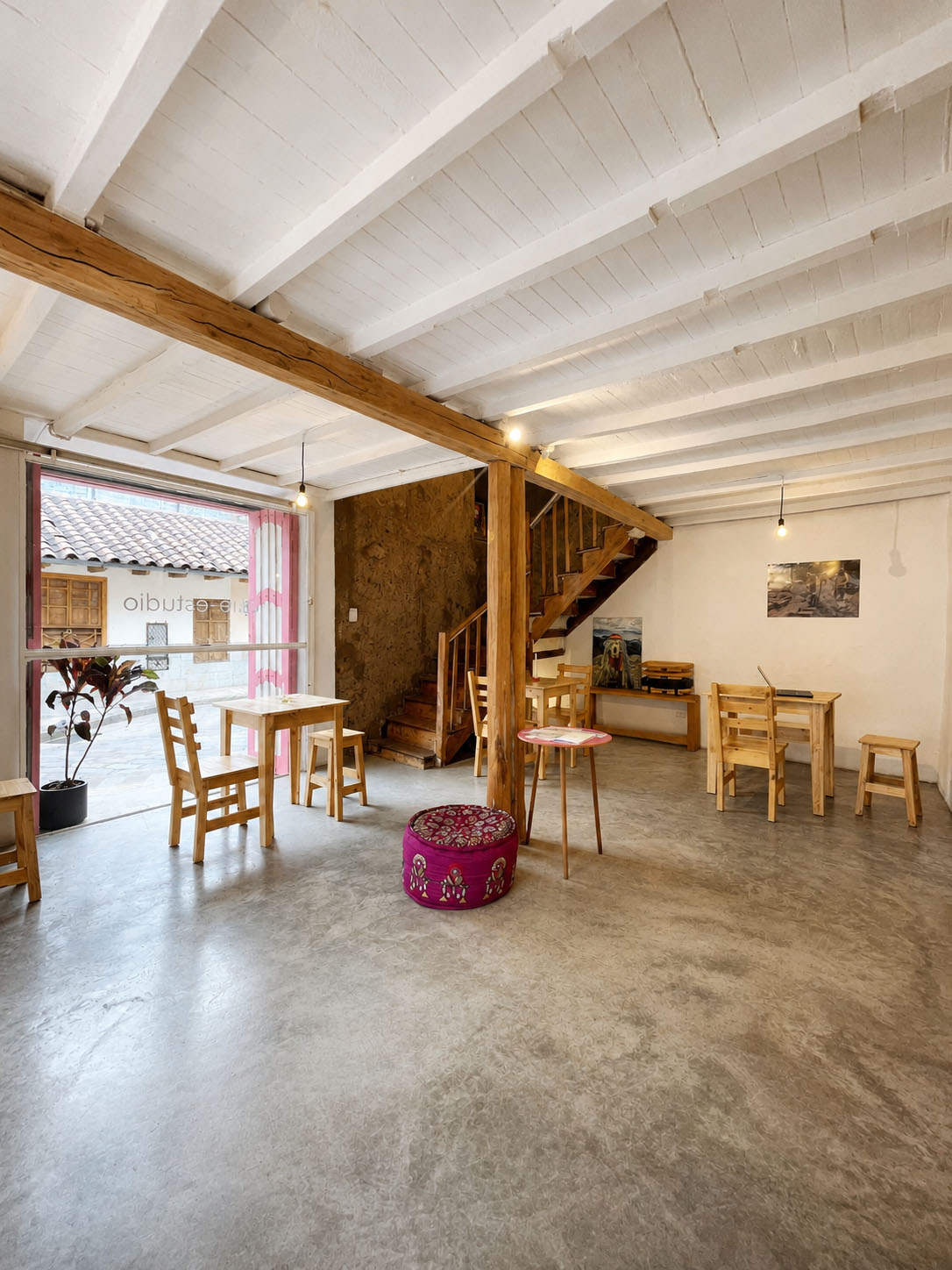 Café-Estudio — el estudio de arquitectura en la planta alta, con tablero de dibujo, escritorio y butaca naranja sobre piso de madera, contra el muro de tapial visto bajo la cubierta blanca