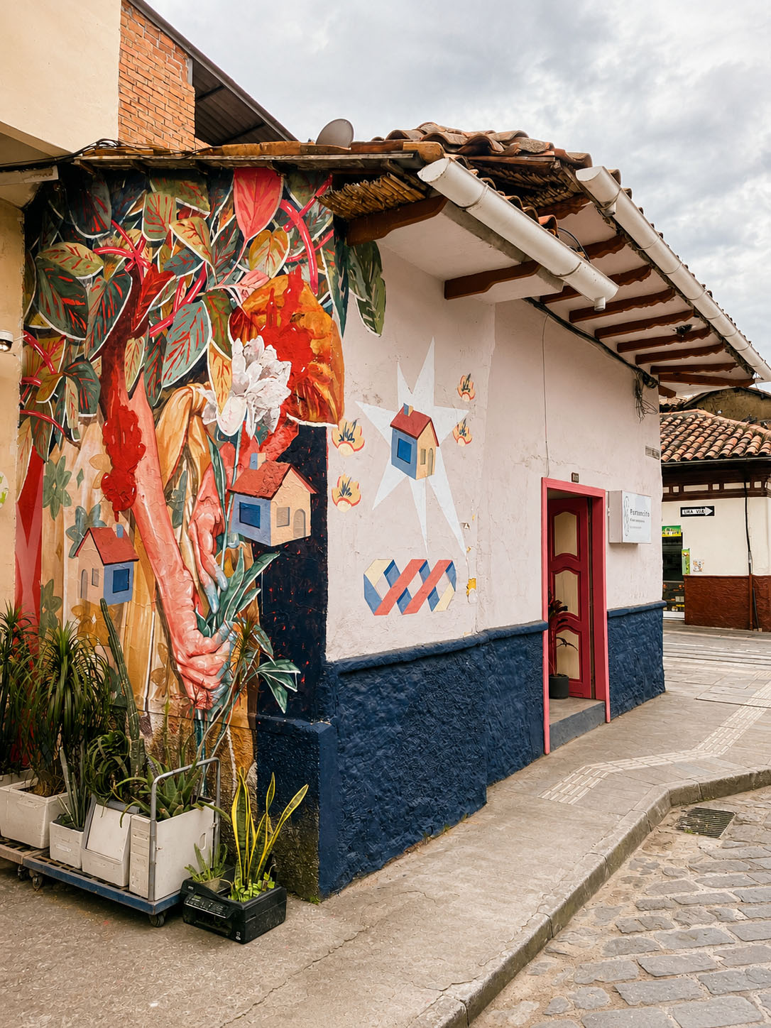 Café-Estudio — salón amplio de mesas de pino con puff fucsia, muro de bahareque visto y puertas plegables rosas abiertas al patio interior