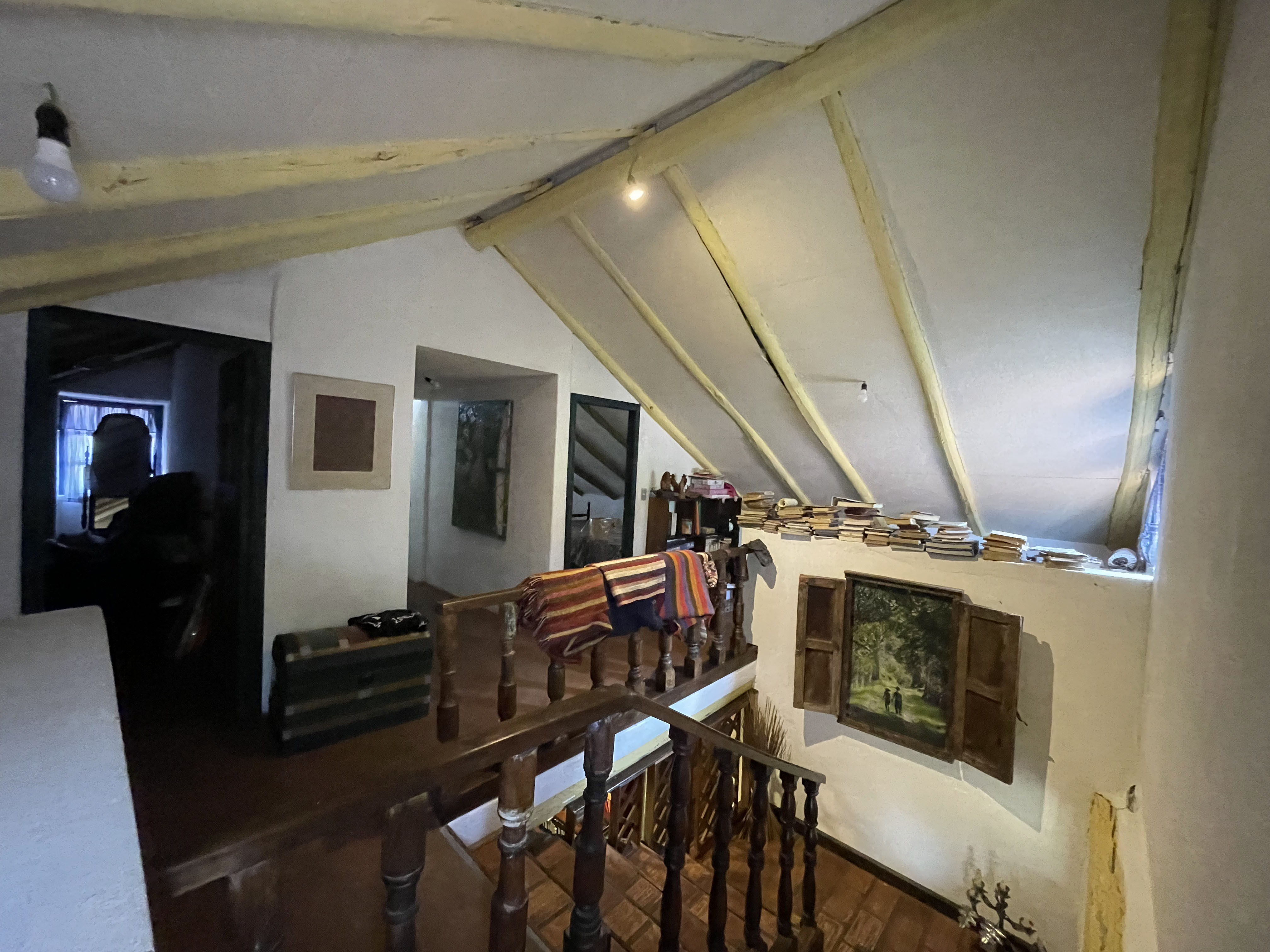Casa de Tapial, antes — desván cerrado bajo la cubierta de madera, penumbra, libros apilados sobre el muro y trastos guardados
