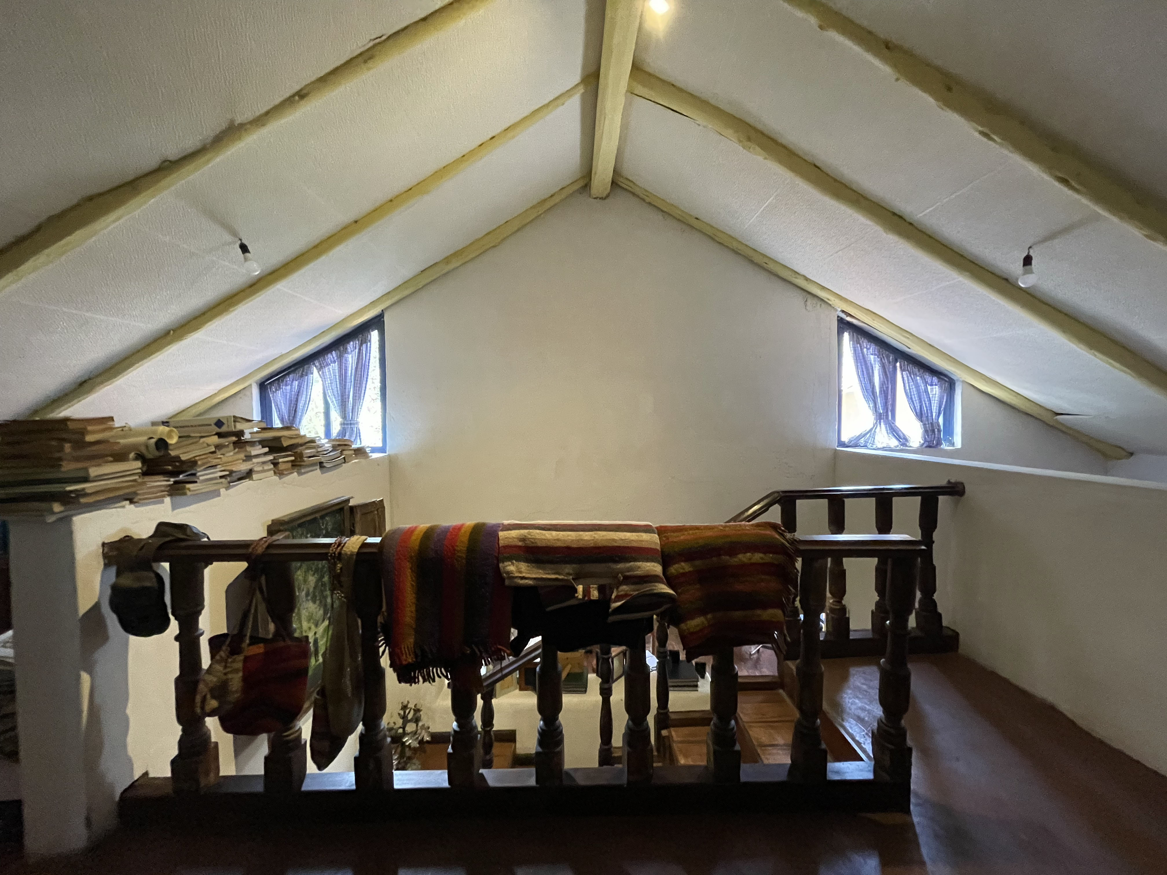 Casa de Tapial, antes — la cumbrera a dos aguas con ventanas pequeñas, vigas pintadas de amarillo y textiles colgados del barandal